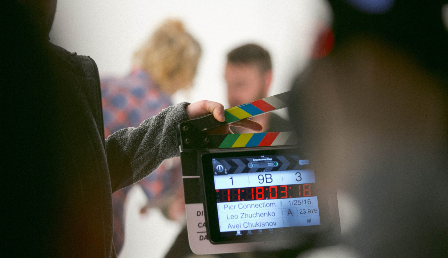 A man holding a clapperboard during a Hollywood movie filming. Actor in the background, blurry, being touched up by the makeup artist using hair thickening fibers.